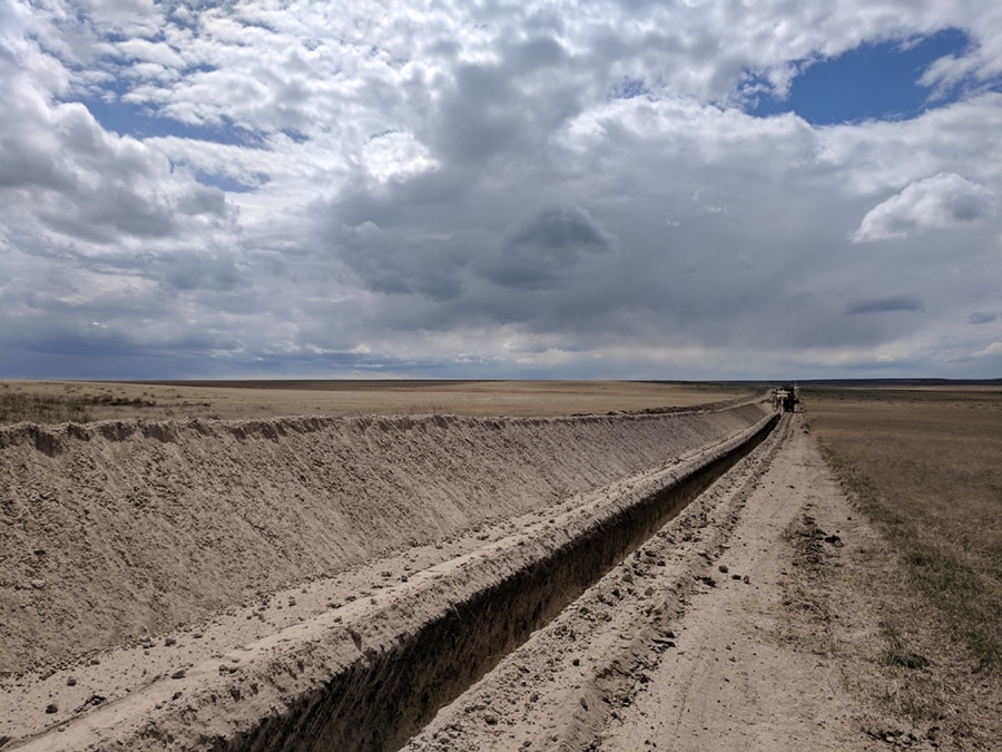 A wide dirt trench is visible in an open landscape, with dry soil on either side. The scene features a cloudy sky above and stretches out to grassy fields in the background.