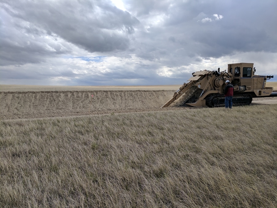 A large heavy machinery vehicle is working on a flat landscape, with a person standing near it for scale. The ground is dry with few grasses, and the sky is filled with clouds, creating a muted atmosphere.