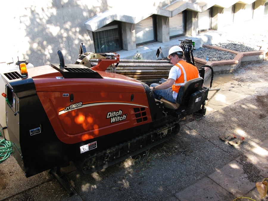 A man in a hard hat and safety vest operates a Ditch Witch JT922 trencher. The machine is positioned on a gravel surface near a building with large windows, and construction materials can be seen in the background.