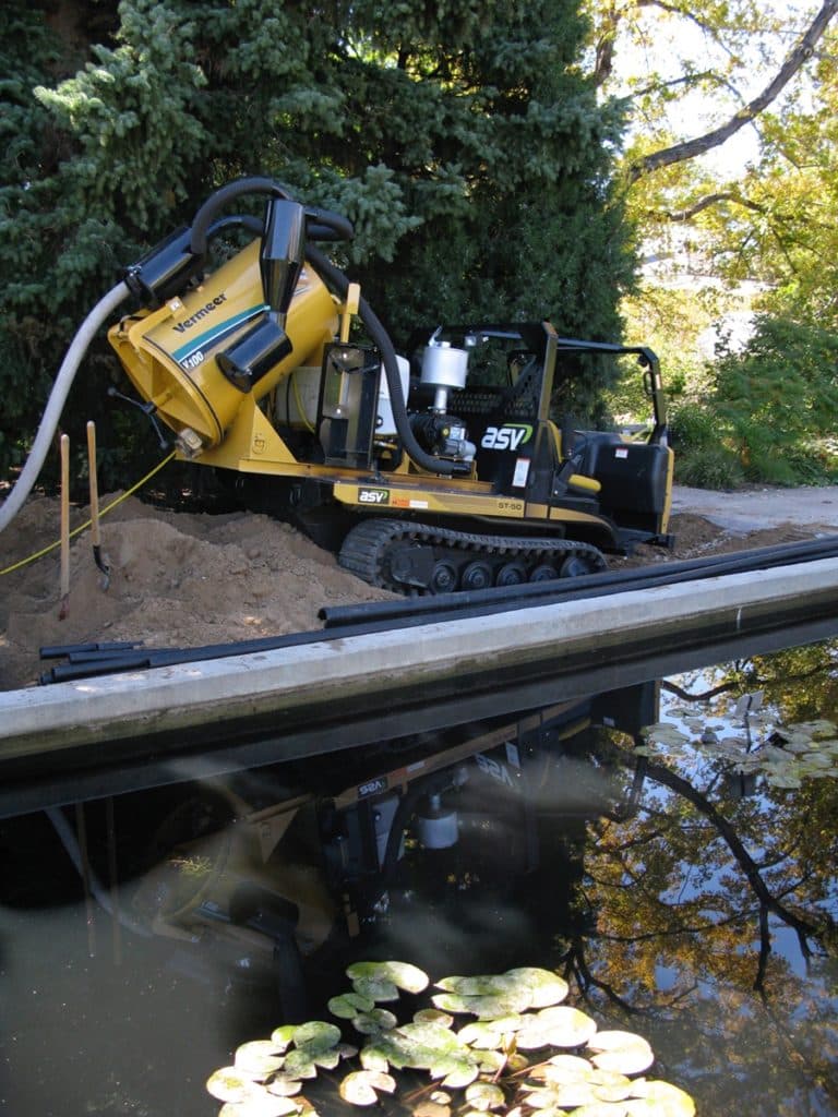 A tracked construction vehicle with a yellow exterior is positioned near a body of water. It is connected to a hose and appears to be working on a pile of earth next to a pond with lily pads visible in the water.