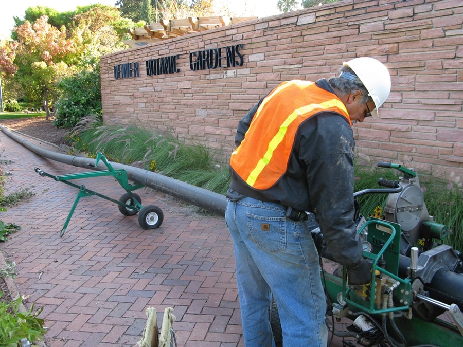 A worker in a construction helmet and orange safety vest is adjusting equipment in front of a sign that reads "Aquatic Gardens." A long tube is connected to a cart behind him, on a brick pathway surrounded by greenery.