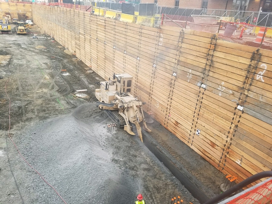 A construction site showing a long wall of wooden sheeting, with a machine digging into the ground below. Various construction equipment and workers in safety gear are visible, as well as piles of gravel on the ground.