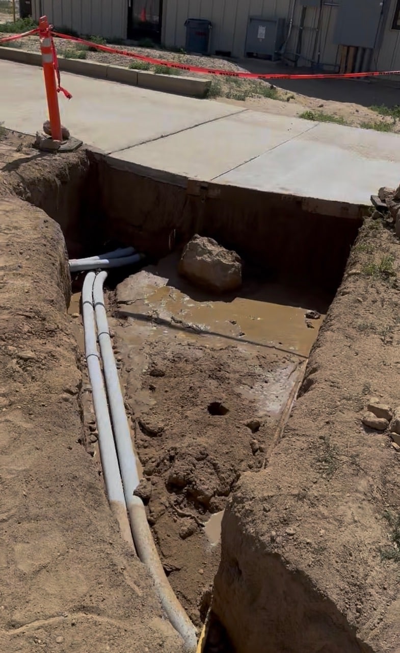 An excavation site shows a rectangular trench filled with water and mud, revealing exposed white pipes and a large rock. In the background, there is a concrete pathway and a red safety cone, alongside a building.