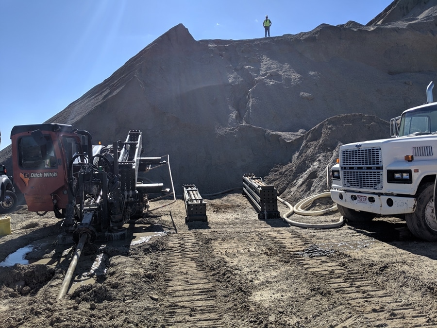 A construction site with a drilling machine positioned on uneven terrain beside a white truck. A person in a green safety vest stands on a dirt mound in the background, and there is a hose and other equipment visible on the ground.