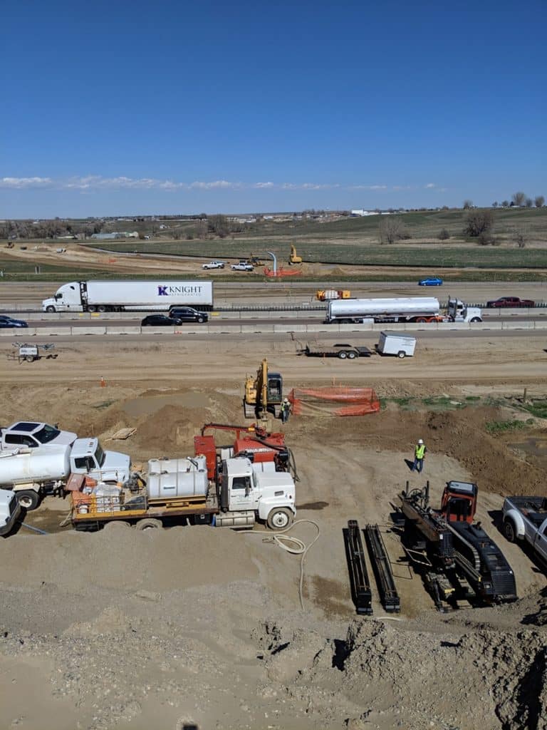 A construction site with heavy machinery and several vehicles in the foreground. In the background, multiple lanes of highway traffic are visible, including trucks and cars, with open fields extending beyond.