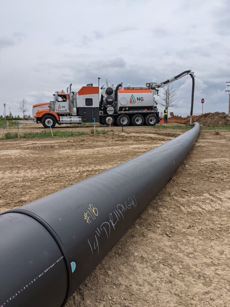 A large black pipe lies flat on the ground, with a construction truck equipped with a vacuum system in the background. The area is an open field with brown earth and cloudy skies overhead.