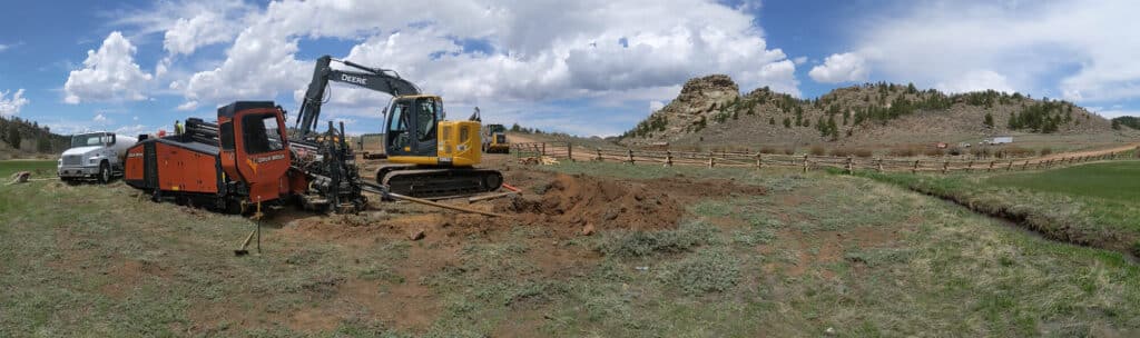 A construction site featuring an orange trencher and a yellow excavator on a grassy landscape. The machinery is positioned near a dug-up area, with distant hills and clouds in the blue sky.