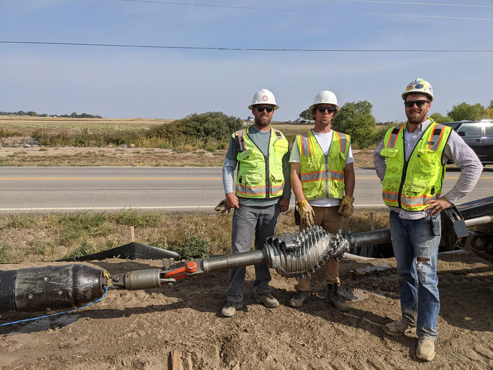 Three construction workers in safety vests and hard hats pose next to a large pipe lying on the ground. The setting includes a rural road, with fields and trees in the background.