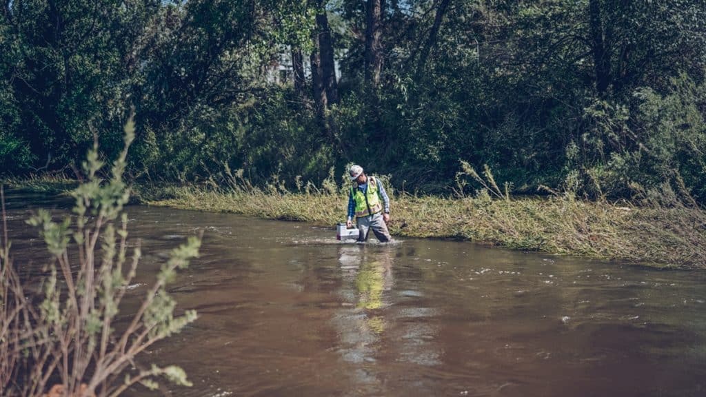 A person wearing a safety vest and a helmet stands in shallow water near a riverbank, holding equipment. The background features dense greenery and trees, with rippling water reflecting light.