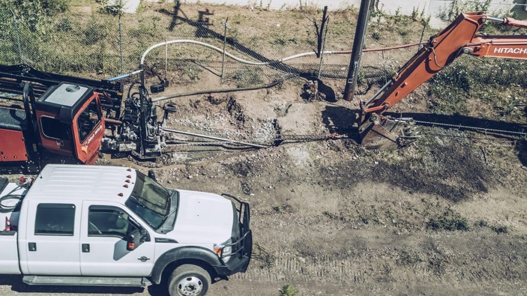 An aerial view of a construction site featuring a white pickup truck next to a large red excavator. The excavator is positioned near a vertical pole, with tools and pipes visible on the ground, and surrounding greenery and a fence in the background.