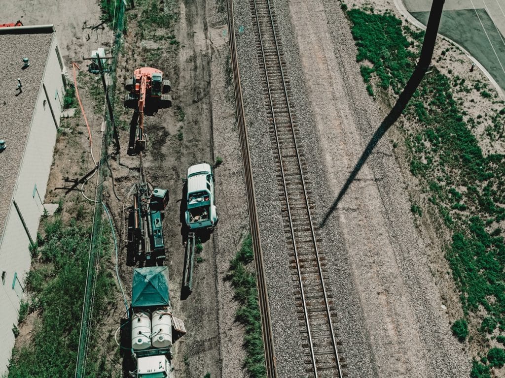 An aerial view of a construction site beside railroad tracks. A white truck is parked near heavy machinery, including a crane and equipment on flatbed trailers, with gravel and dirt along the tracks. The surrounding area shows green grass and a building.