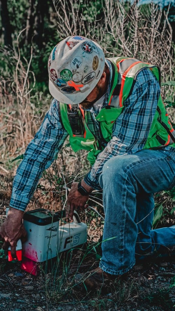 A man wearing a safety helmet and a green high-visibility vest kneels beside a white device on the ground. He is adjusting the device while surrounded by tall grass and trees in the background.