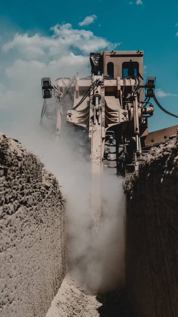 A heavy construction vehicle is operating in a trench, creating dust clouds as it moves. The scene features a blue sky with scattered clouds in the background, highlighting the construction activity.