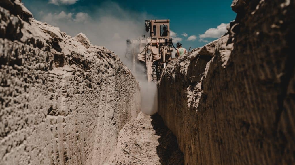 A construction machine operates in a deep trench, creating dust as it moves. The trench walls are uneven, showing freshly dug earth, while a blue sky with scattered clouds is visible above.