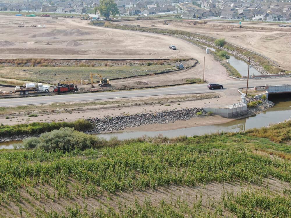 A wide view of a construction site near a riverbank. Trucks and construction equipment are visible on a roadway, with a rocky riverbank and patches of vegetation in the foreground. The background features undeveloped land and residential buildings.