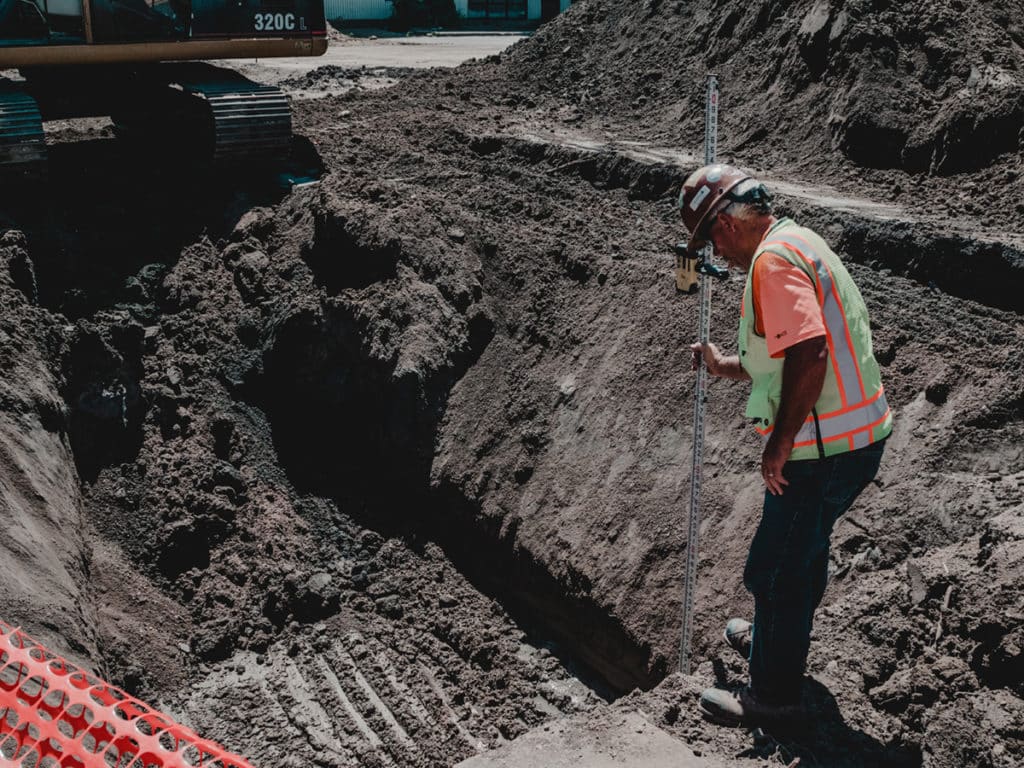 A construction worker in a safety vest examines a deep trench in the ground, holding a measuring rod. An excavator is visible in the background, with piles of dirt surrounding the site.