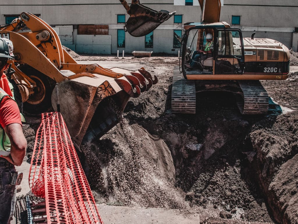 Two heavy construction machines are operating near a deep excavation site. One machine, a backhoe, is digging into the ground, while an excavator is positioned nearby, both surrounded by orange safety fencing and a partially visible building in the background.